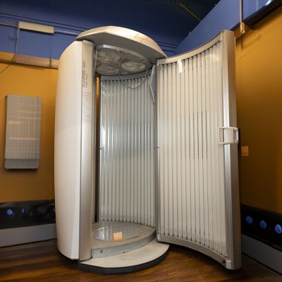 A vertical tanning booth in a room with yellow and blue walls, featuring open doors revealing white interior panels and wood flooring.