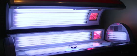 Open tanning bed with glowing fluorescent bulbs, emitting a blue and white light. Red-tinted areas on the sides suggest heat lamps. A small sign is visible.