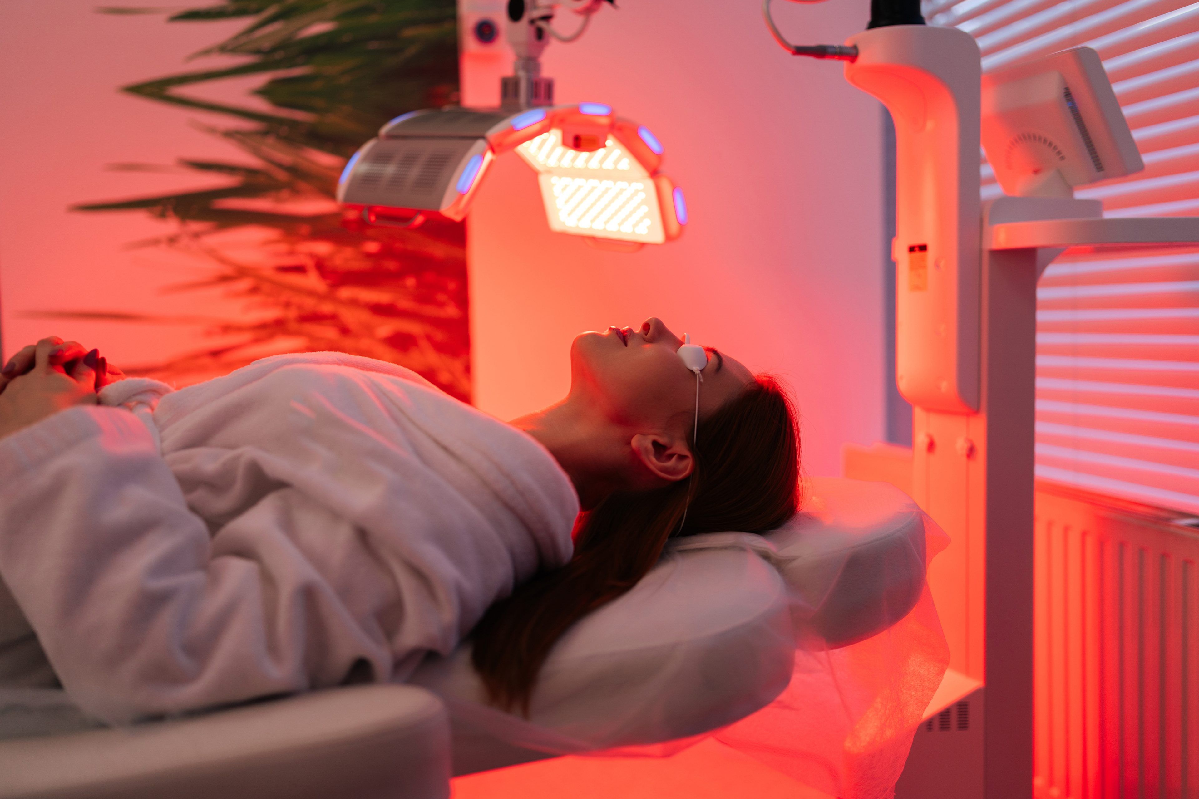 A person in a spa robe lies on a treatment table under red LED light therapy. The room is softly lit and calming, creating a relaxing atmosphere.