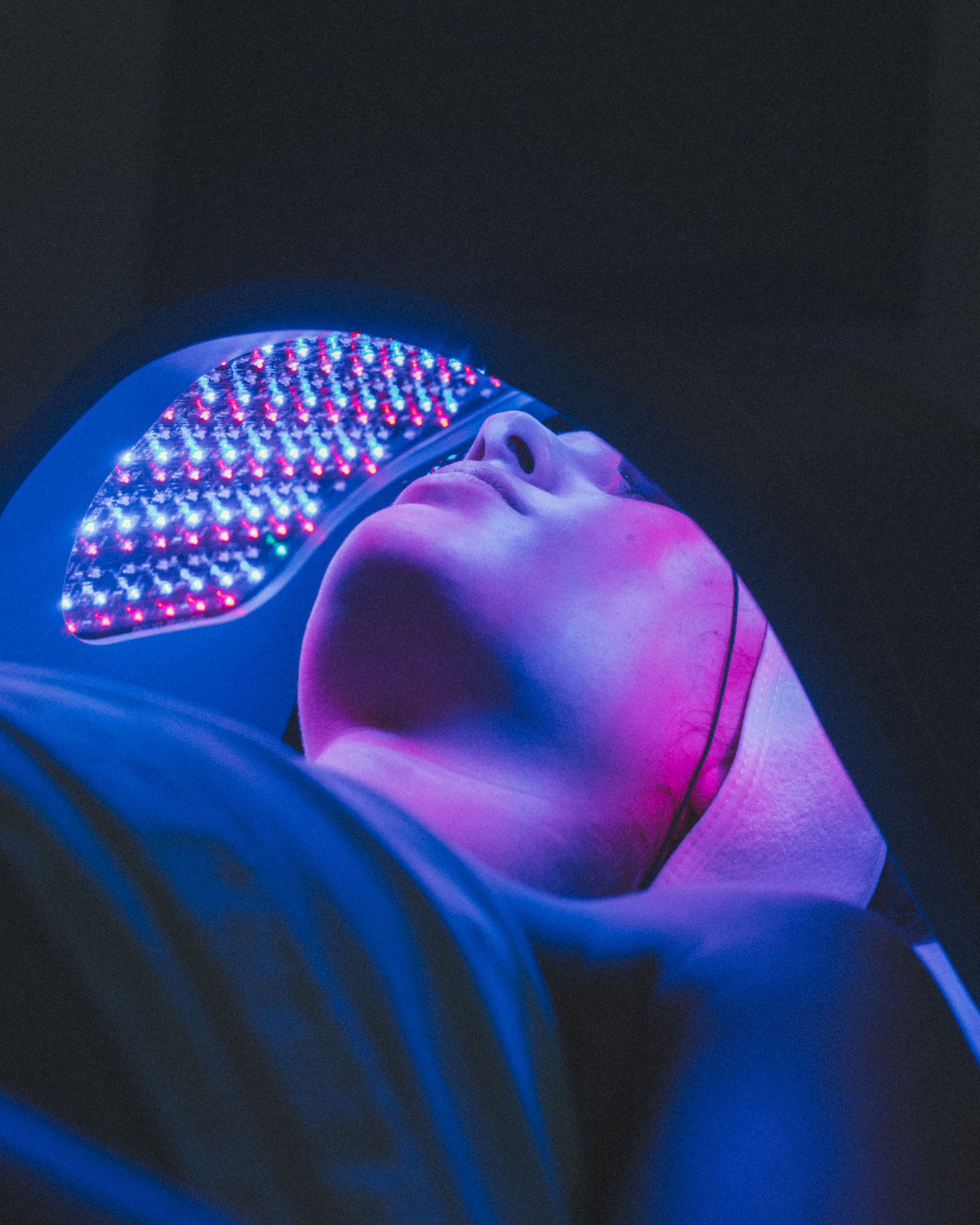 A person is lying under a glowing LED therapy mask, emitting blue and pink light in a dark setting, creating a serene and futuristic atmosphere.