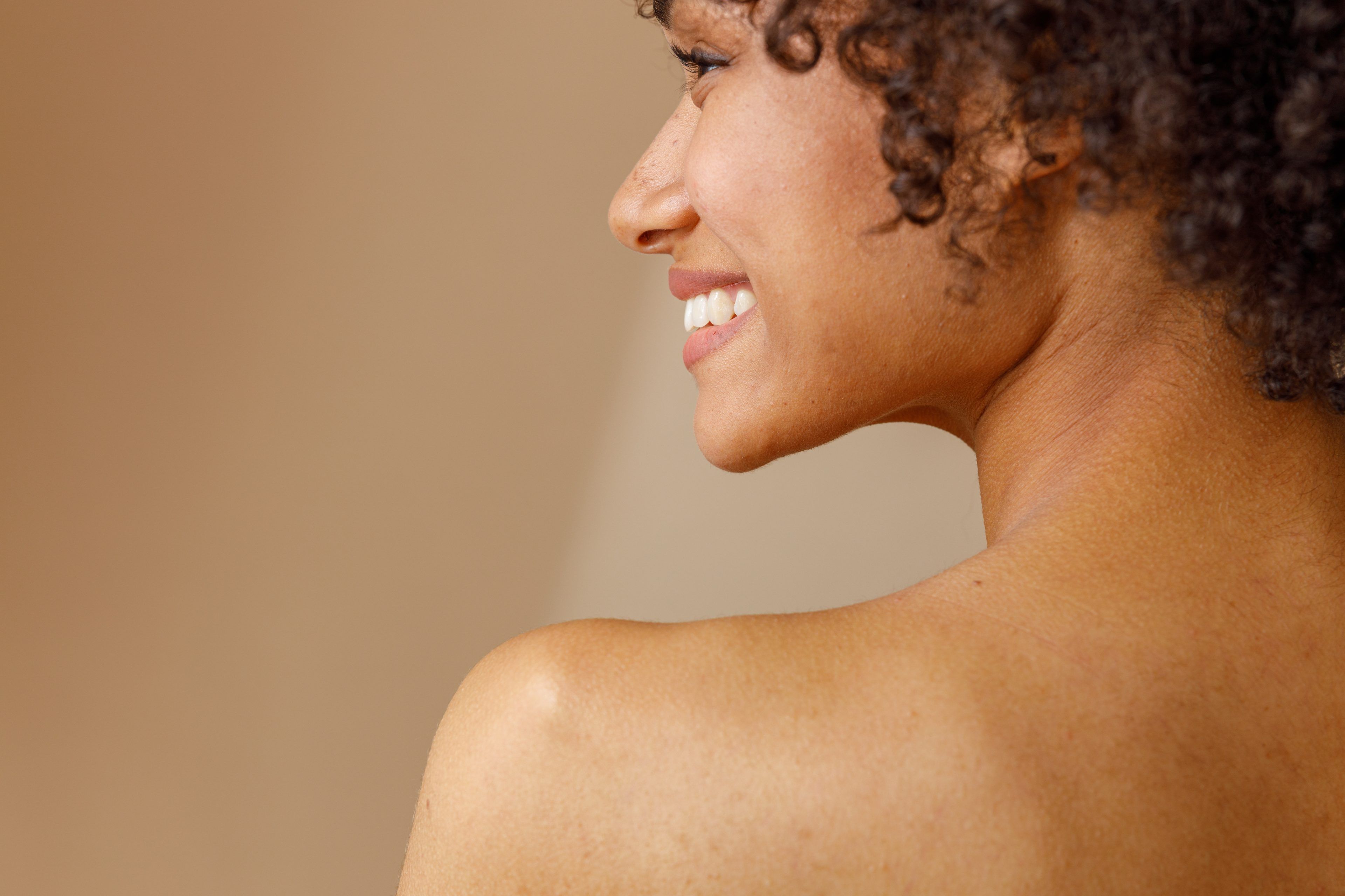 A woman with curly hair smiles warmly, facing away from the camera over her bare shoulder. The background is a soft beige, conveying a calm and natural tone.