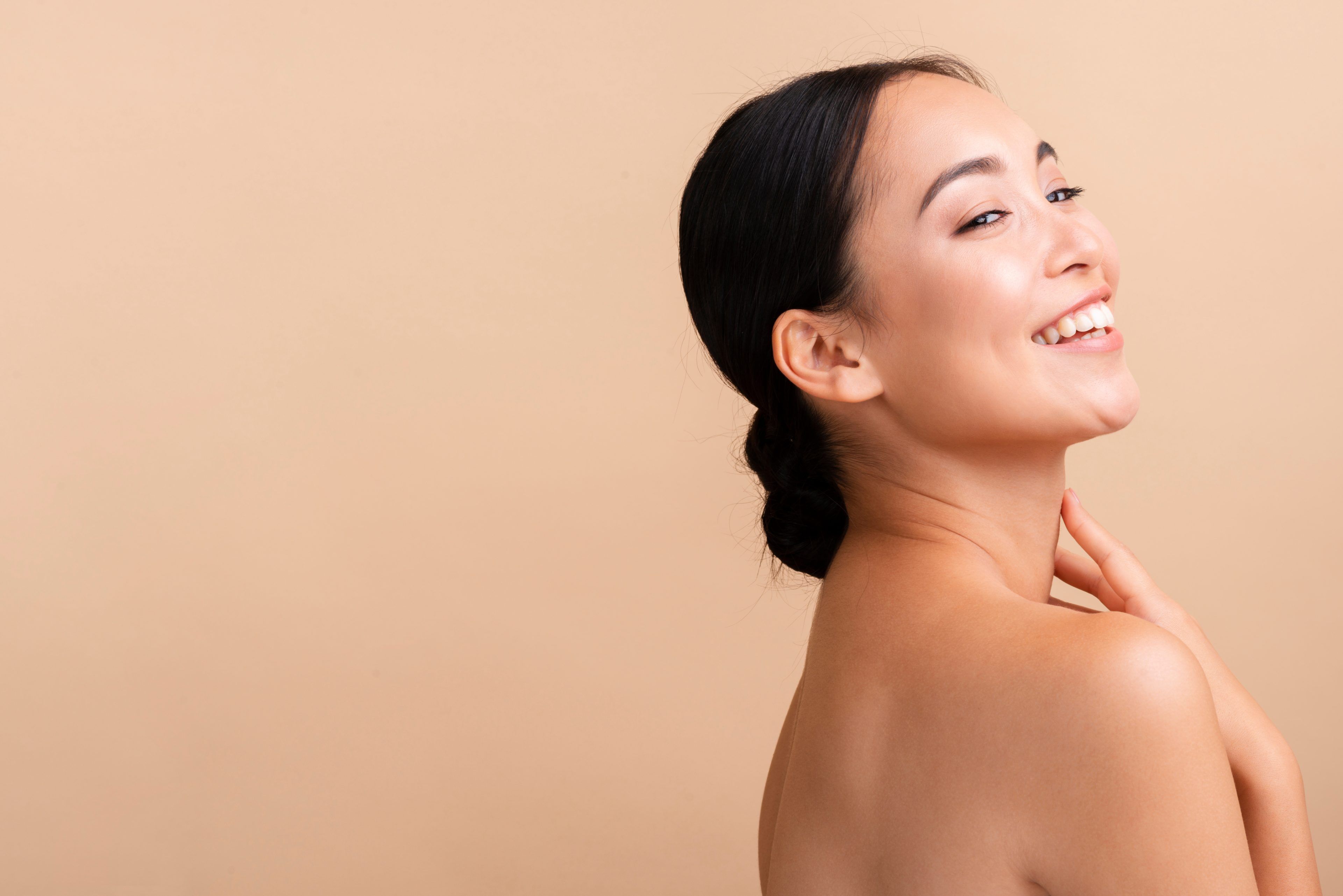 Smiling woman with dark hair in a bun, turned sideways against a soft beige background. She exudes a sense of joy and natural beauty.