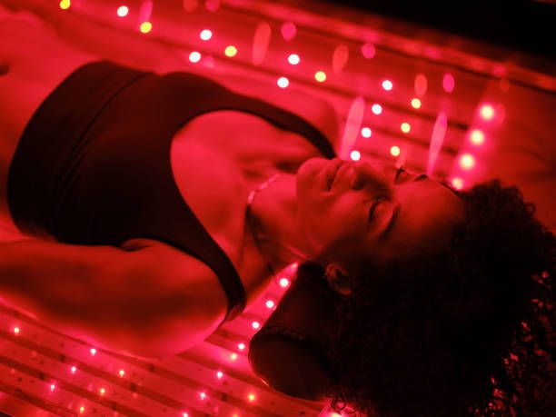 Woman in a black sports bra relaxes on a surface illuminated by soft red LED lights, eyes closed, conveying a sense of calm and rejuvenation.