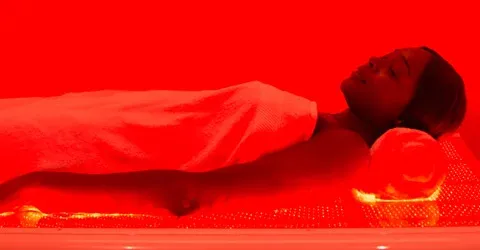 A person relaxes on a glowing red light therapy bed, covered with a towel. The scene emits a warm, calming atmosphere, suggesting relaxation and wellness.