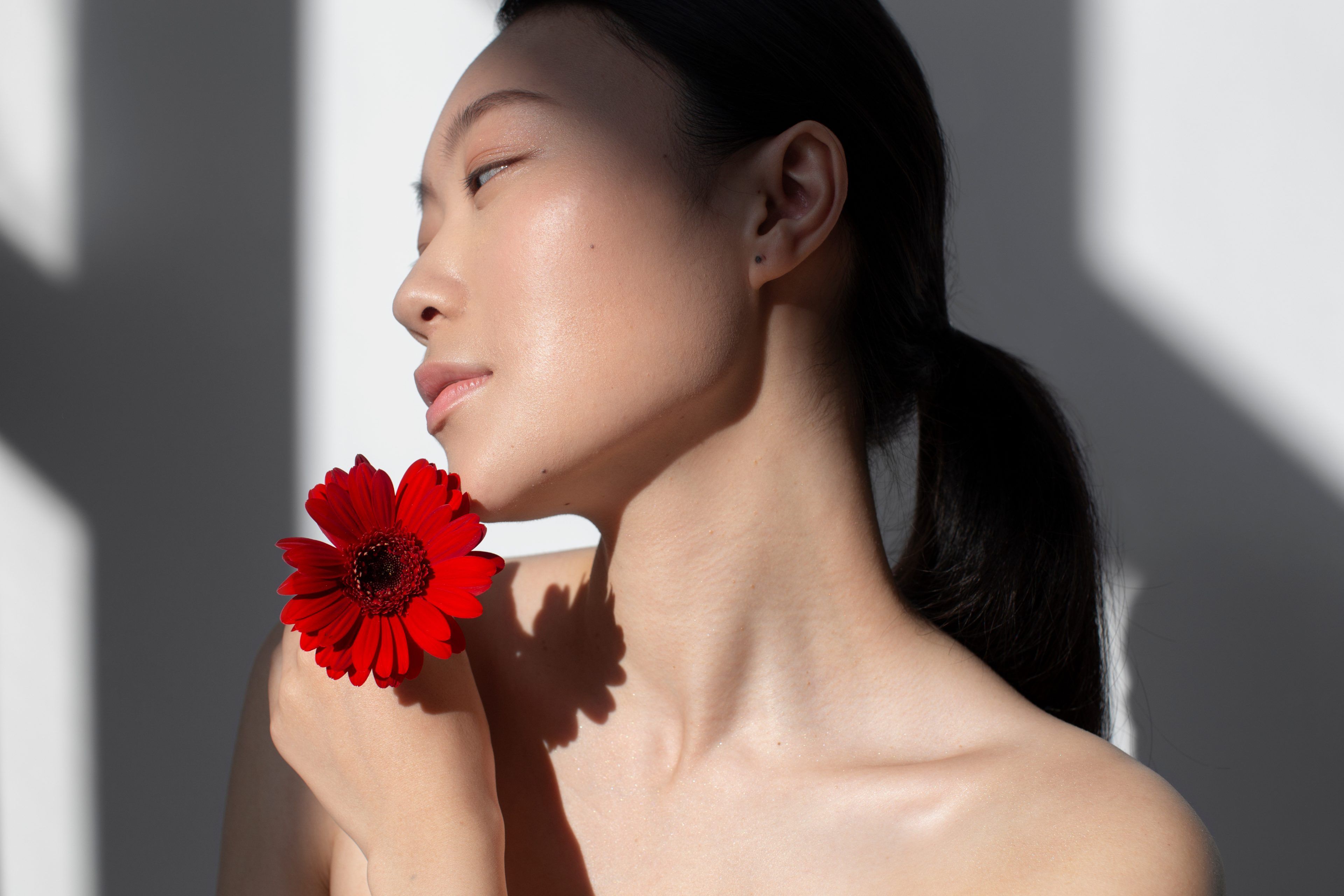 A woman with smooth skin poses gracefully in soft light. She holds a vibrant red flower near her shoulder, conveying elegance and tranquility.