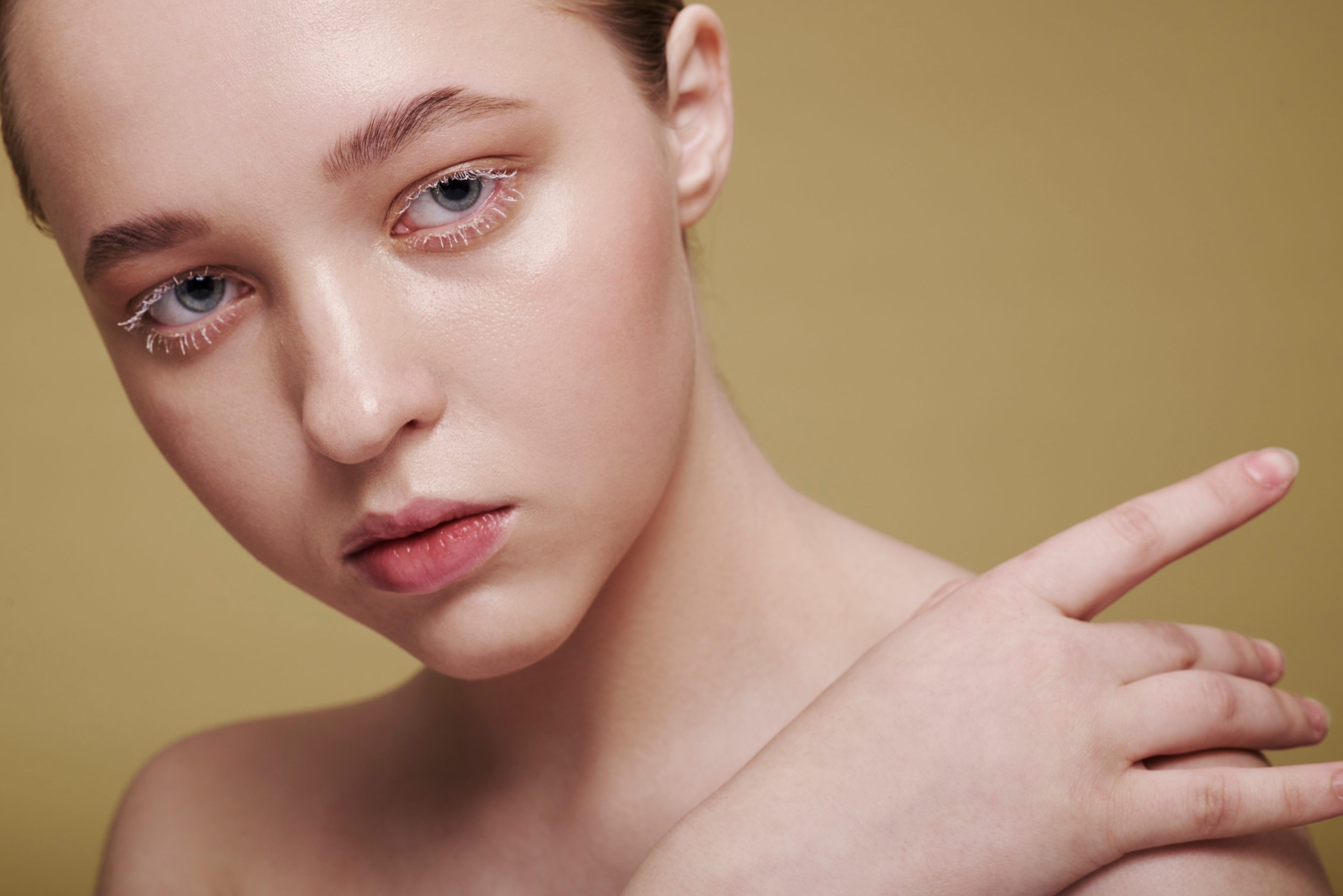 Close-up of a person with light eyeshadow and white mascara, looking thoughtfully into the distance. The background is a soft beige, creating a serene tone.